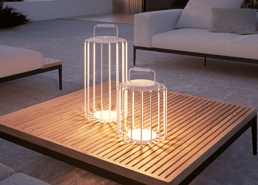 Two modern white lanterns with handles sit on a wooden slatted coffee table in an outdoor seating area with sofas at dusk.